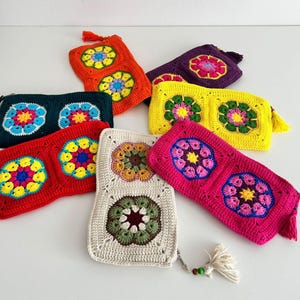 May include: A collection of colorful, handmade crochet pouches. Each pouch features a floral design in various colors, including orange, yellow, red, green, and pink. The pouches have a rectangular shape and are adorned with tassels.