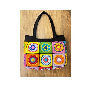 May include: A colorful crocheted tote bag with black straps. The bag is made of squares with different colored flowers in the center.