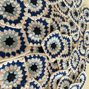 Puede incluir: Primer plano de una pila de cuadrados tejidos a crochet en tonos de azul, blanco y negro. Los cuadrados tienen un patrón de estrella en el centro.