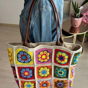 May include: A large, colorful crocheted tote bag with a brown leather handle. The bag is made of squares with a flower design in shades of pink, yellow, blue, green, orange, and red.