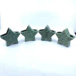 May include: Four green star-shaped ceramic candle holders spelling "NOEL". Each star has a different letter and is decorated with a leaf and berry pattern. The candle holders are a festive holiday decoration.
