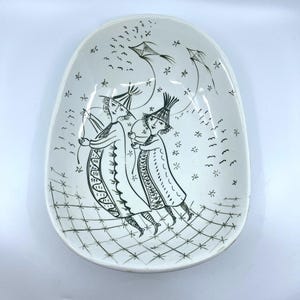 May include: White oval ceramic serving dish with a black illustration of two figures with kites and birds. The figures are wearing long coats and hats. The dish has a grid pattern on the bottom.