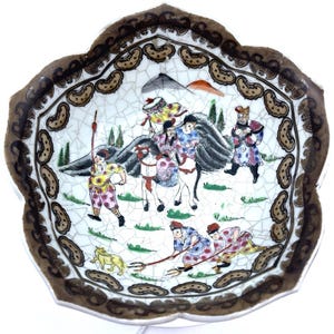 May include: A white porcelain bowl with a brown and gold rim. The bowl features a hand-painted scene of people on horseback and other figures in a mountainous landscape. The bowl has a crackled glaze.