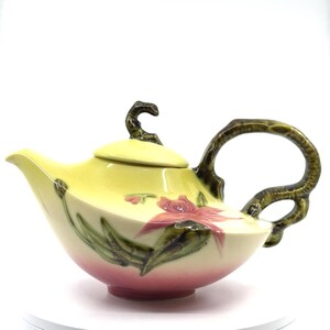 May include: A vintage ceramic teapot with a yellow and pink gradient. The teapot features a floral design with red flowers and green leaves. The handle and lid handle are dark green. The teapot is a decorative item.