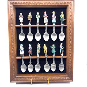 May include: A framed collection of decorative spoons. Each spoon has a detailed figurine at the top, depicting various people in traditional attire. The spoons are displayed in a wooden frame with a dark velvet backing.