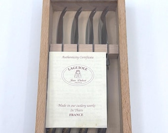 Könnte beinhalten: Ein Set aus vier silberfarbenen Käsemessern mit gebogenen Spitzen, untergebracht in einer hellen Holzschachtel. Über den Messern liegt ein Echtheitszertifikat mit dem Text: Authenticity Certificate, LAGUIOLE, Jean Dubost, FRANCE, Made in our cutlery works, In Thiers, FRANCE.