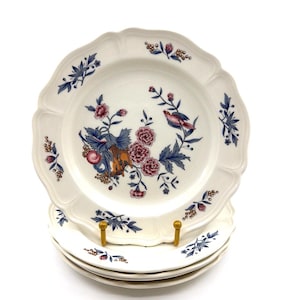 May include: A stack of cream-colored ceramic plates with scalloped edges. The plates feature a central botanical pattern with pink flowers, blue leaves, and a small bird, complemented by smaller floral sprigs along the scalloped rim.