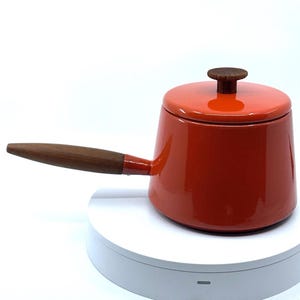 May include: A small, red enameled saucepan with a wooden handle and lid. The lid has a wooden knob. The saucepan is on a white surface. The handle is approximately 7 inches long.