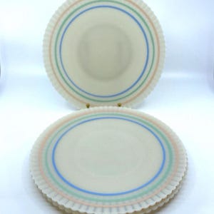 May include: Off-white plates with scalloped edges and three concentric rings in pastel colors: pink, green, and blue. The plates are stacked and arranged on a white surface, showcasing a vintage aesthetic.