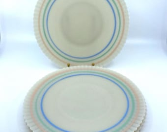 May include: Off-white plates with scalloped edges and three concentric rings in pastel colors: pink, green, and blue. The plates are stacked and arranged on a white surface, showcasing a vintage aesthetic.