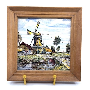 May include: A framed tile featuring a Dutch windmill scene with a bridge and water. The wooden frame has two gold-colored hooks. The artwork depicts a rural landscape with a windmill, houses, and a bridge over a river.