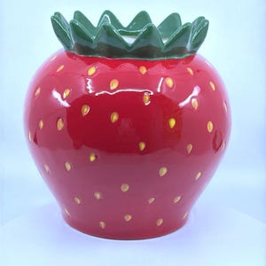 May include: A ceramic strawberry-shaped container with a glossy red finish. The container features yellow seed details and a green leafy top. This decorative piece is ideal for kitchen or home decor.