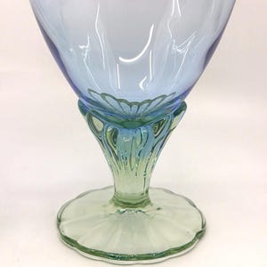 K&ouml;nnte beinhalten: Ein dekorativer Glasbecher mit einer hellblauen Schale und einem gr&uuml;nen Stiel und Fu&szlig;. Der Becher hat ein Blumendesign am Boden der Schale und einen gewellten Rand am Fu&szlig;. Das Glas ist transparent.