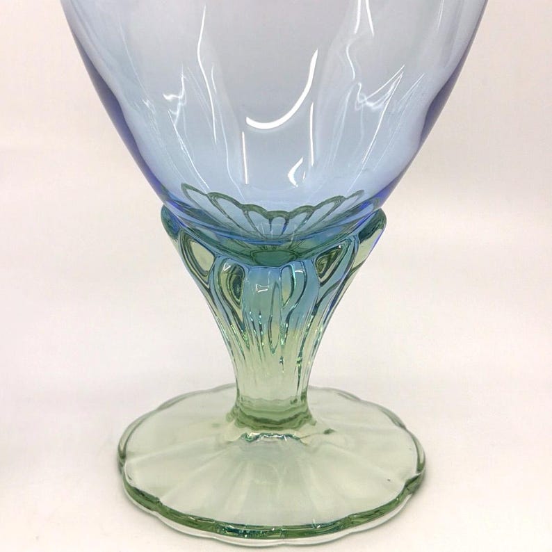 K&ouml;nnte beinhalten: Ein dekorativer Glasbecher mit einer hellblauen Schale und einem gr&uuml;nen Stiel und Fu&szlig;. Der Becher hat ein Blumendesign am Boden der Schale und einen gewellten Rand am Fu&szlig;. Das Glas ist transparent.