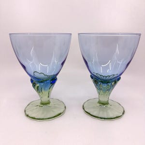 K&ouml;nnte beinhalten: Zwei elegante blaue Weingl&auml;ser mit dekorativen Stielen und F&uuml;&szlig;en. Die Kelche sind hellblau, gehen in dunkleres Blau und Gr&uuml;n an den Stielen &uuml;ber. Die F&uuml;&szlig;e sind hellgr&uuml;n und bilden einen auff&auml;lligen Kontrast.