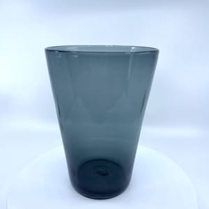 May include: A tall, cylindrical, smoky-blue glass vase. The vase has a slightly flared top and a rounded base. The glass is smooth and transparent, allowing light to pass through. The vase is empty and sits on a white surface.