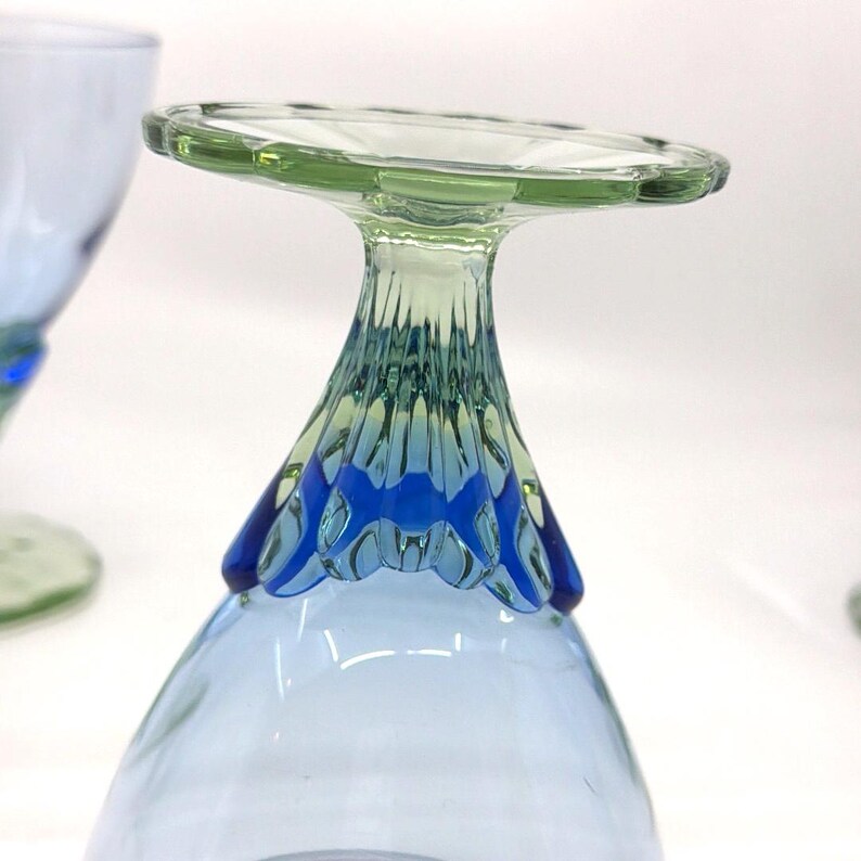 K&ouml;nnte beinhalten: Ein hellblaues Glasbecher mit gr&uuml;nem Rand und blauem Zierfu&szlig;. Der Stiel hat ein strukturiertes Design. Der Becher ist umgedreht, wodurch die Details des Fu&szlig;es und des Rands sichtbar werden.