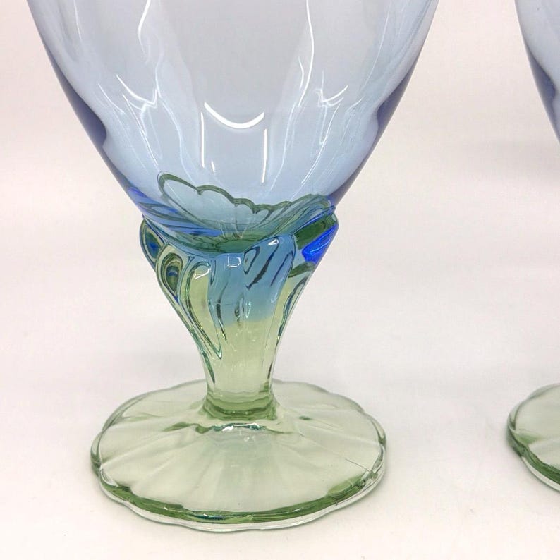 K&ouml;nnte beinhalten: Zwei Weingl&auml;ser mit blau-gr&uuml;nem Stiel. Die Schale des Glases ist hellblau und geht am Rand in ein dunkleres Blau &uuml;ber. Der Stiel hat ein Blumendesign mit gr&uuml;nen und blauen Akzenten, und der Fu&szlig; ist gr&uuml;n und gewellt.
