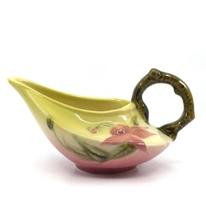 May include: A vintage ceramic gravy boat with a yellow interior and a pink exterior. The boat features a floral design with pink flowers and green leaves. The handle is brown and has a branch-like design.