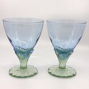 K&ouml;nnte beinhalten: Zwei blau get&ouml;nte Weingl&auml;ser mit einem einzigartigen Design. Die Kelche sind hellblau, am Boden dunkler. Die Stiele weisen ein Blumendesign in Gr&uuml;n und Blau auf, das auf einem gr&uuml;nen kreisf&ouml;rmigen Sockel ruht.