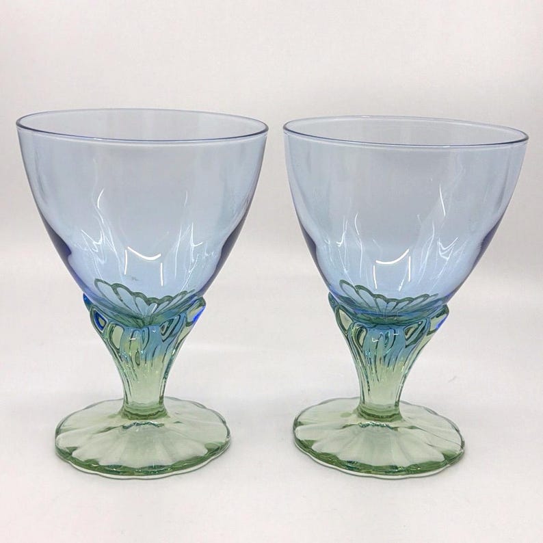K&ouml;nnte beinhalten: Zwei blau get&ouml;nte Weingl&auml;ser mit einem einzigartigen Design. Die Kelche sind hellblau, am Boden dunkler. Die Stiele weisen ein Blumendesign in Gr&uuml;n und Blau auf, das auf einem gr&uuml;nen kreisf&ouml;rmigen Sockel ruht.