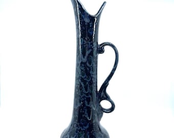 May include: A tall, dark blue ceramic vase with a unique handle and a flared base. The vase has a speckled pattern in shades of blue and white. The vase is approximately 12 inches tall.
