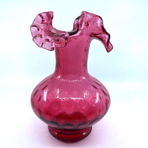 May include: A ruby red glass vase with a ruffled top and a textured, bubbled design. The vase has a round body and a flared base.