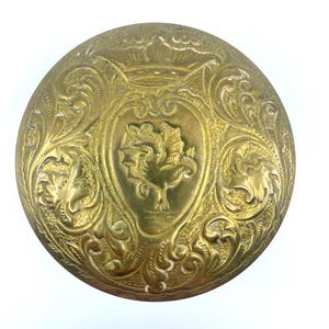 May include: A round, gold-colored metal button with an intricate, engraved design. The center of the button features a stylized bird or animal with a heart-shaped frame.
