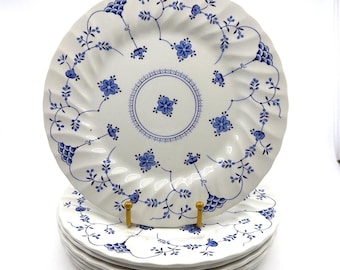 Könnte beinhalten: Ein Stapel weißer Keramikteller mit einem blauen Blumenmuster aus zarten Ranken und kleinen Blüten. Die Teller haben einen gewellten, geschwungenen Rand und ein zentrales kreisförmiges Motiv mit einer blauen Blume und einer gepunkteten Umrandung.
