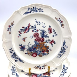 May include: A cream-colored ceramic plate with a scalloped edge, featuring a central floral motif in shades of blue, red, and orange. The design includes a small bird, a vase, and fruit, complemented by delicate blue leaf sprigs along the rim.