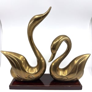 May include: A pair of polished brass swan figurines mounted on a rectangular dark wood base. The left swan has an elongated neck reaching upward, while the right swan has a curved neck facing downward. Both figures feature smooth, reflective surfaces.