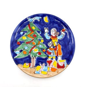 May include: A round, hand-painted ceramic plate with a Christmas theme. The plate features a dark blue background with a green Christmas tree, figures, presents, and a crescent moon. The plate has a light brown rim.