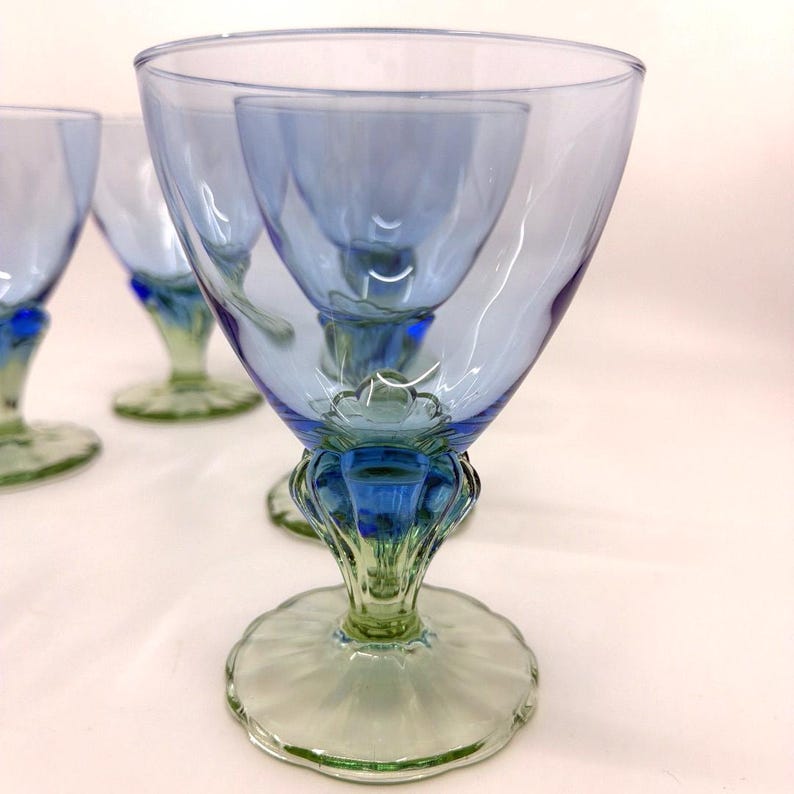 K&ouml;nnte beinhalten: Satz von vier blau-gr&uuml;nen Weingl&auml;sern. Die Gl&auml;ser haben eine ausgestellte Schalenform mit blauem Farbton, einen detaillierten blauen Stiel und einen gr&uuml;nen Fu&szlig;. Die Gl&auml;ser sind auf einer wei&szlig;en Oberfl&auml;che angeordnet.