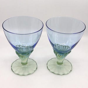 K&ouml;nnte beinhalten: Zwei blau get&ouml;nte Weingl&auml;ser mit einzigartigem Design. Die Kelche sind hellblau mit einem dunkleren blauen Rand. Die Stiele sind gr&uuml;n mit einem dekorativen Blumendesign. Der Fu&szlig; ist hellgr&uuml;n mit einem gewellten Rand. Die Gl&auml;ser sind leer.