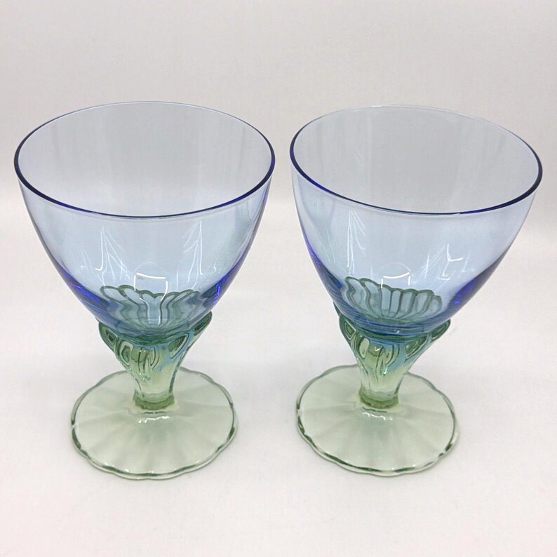 K&ouml;nnte beinhalten: Zwei blau get&ouml;nte Weingl&auml;ser mit einzigartigem Design. Die Kelche sind hellblau mit einem dunkleren blauen Rand. Die Stiele sind gr&uuml;n mit einem dekorativen Blumendesign. Der Fu&szlig; ist hellgr&uuml;n mit einem gewellten Rand. Die Gl&auml;ser sind leer.