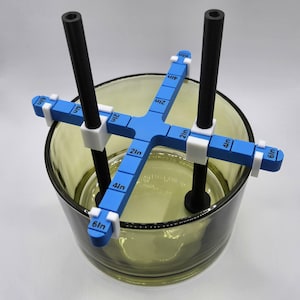May include: A blue and white candle making tool with two black rods and a clear glass jar. The tool has markings in inches from 1 to 6.