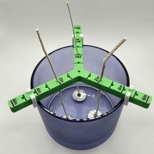 May include: Green candle wick centering tool with three arms, each marked with inch measurements from 1 to 6 inches. The tool is holding three wicks in a clear glass jar with a purple tint.