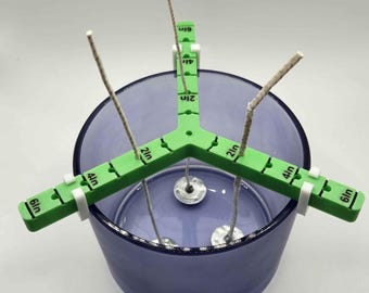 May include: Green candle wick centering tool with three arms, each marked with inch measurements from 1 to 6 inches. The tool is holding three wicks in a clear glass jar with a purple tint.