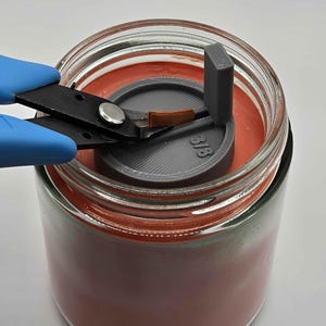 May include: A gray 3D printed candle holder with a wick and a small piece of copper wire being cut with blue handled wire cutters. The holder is sitting inside a clear glass jar with a red candle.