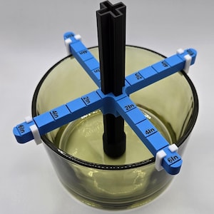 May include: A blue and black floral arranging tool with measurements in inches. The tool is placed inside a clear, olive-green glass bowl. The tool has a central black post with four blue arms extending outwards, each marked with measurements.