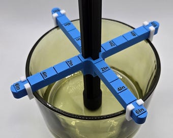 May include: A blue and black floral arranging tool with measurements in inches. The tool is placed inside a clear, olive-green glass bowl. The tool has a central black post with four blue arms extending outwards, each marked with measurements.