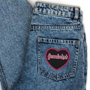 Puede incluir: Un par de pantalones vaqueros azules con un parche negro en forma de corazón que dice "feminista" en letras rosas.