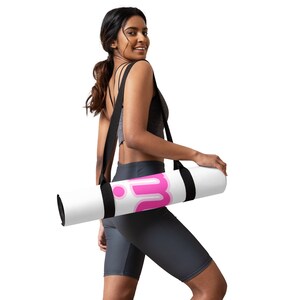 Puede incluir: Una esterilla de yoga blanca con un logotipo "W" rosa y una correa de transporte negra. La esterilla está enrollada y la sostiene una persona.