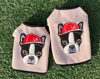 May include: Two pink fleece dog vests with black trim. Each vest features an embroidered French bulldog wearing a red baseball cap with the letters "LA" on it.