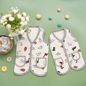 May include: Two off-white baby vests with a dinosaur pattern and gray trim. Each vest has a pocket and button closure. The vests are displayed on a green surface with decorative elements, including flowers and colorful pom-poms.