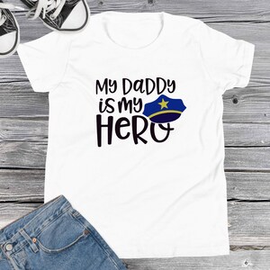 Puede incluir: Camiseta blanca con el texto "My Daddy is my Hero" y un gorro azul de policía con una estrella dorada en la parte superior.
