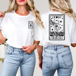 Op de afbeelding: Wit T-shirt met een zwarte afbeelding op de voor- en achterkant. De afbeelding aan de voorkant is een handgebaar in een ruitvorm. De afbeelding op de achterkant toont een handgebaar met nagelgereedschap, rozen en de tekst "THE NAIL TECH".