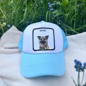 May include: A light blue trucker hat with a white front panel featuring a dog portrait and the name "MOOSE". The hat has a curved brim and a mesh back, perfect for sunny days.