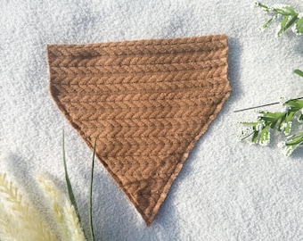 May include: A brown, triangular pet bandana with a textured, woven pattern. The bandana is made of a soft material and is designed to be worn around a pet's neck. The background is a white, textured surface with some greenery.