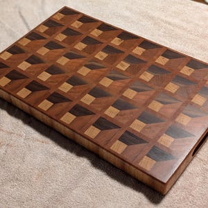 Könnte beinhalten: Ein rechteckiges Schneidebrett aus Holz mit Hirnholzkonstruktion und geometrischem Muster. Die Oberfläche zeigt ein sich wiederholendes Design aus hellbraunen, mittelbraunen und dunkelbraunen Holzblöcken, die einen 3D-Effekt erzeugen.